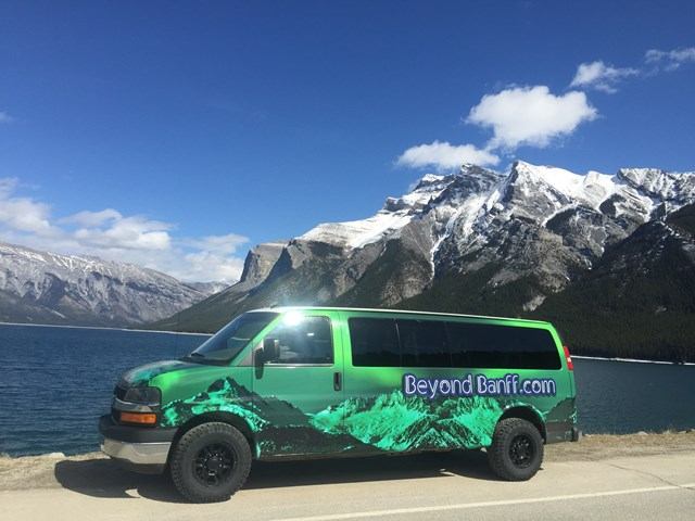 Green Cube Beyond Banff van parked by scenic lake, towering mountains behind under a partly cloudy sky.