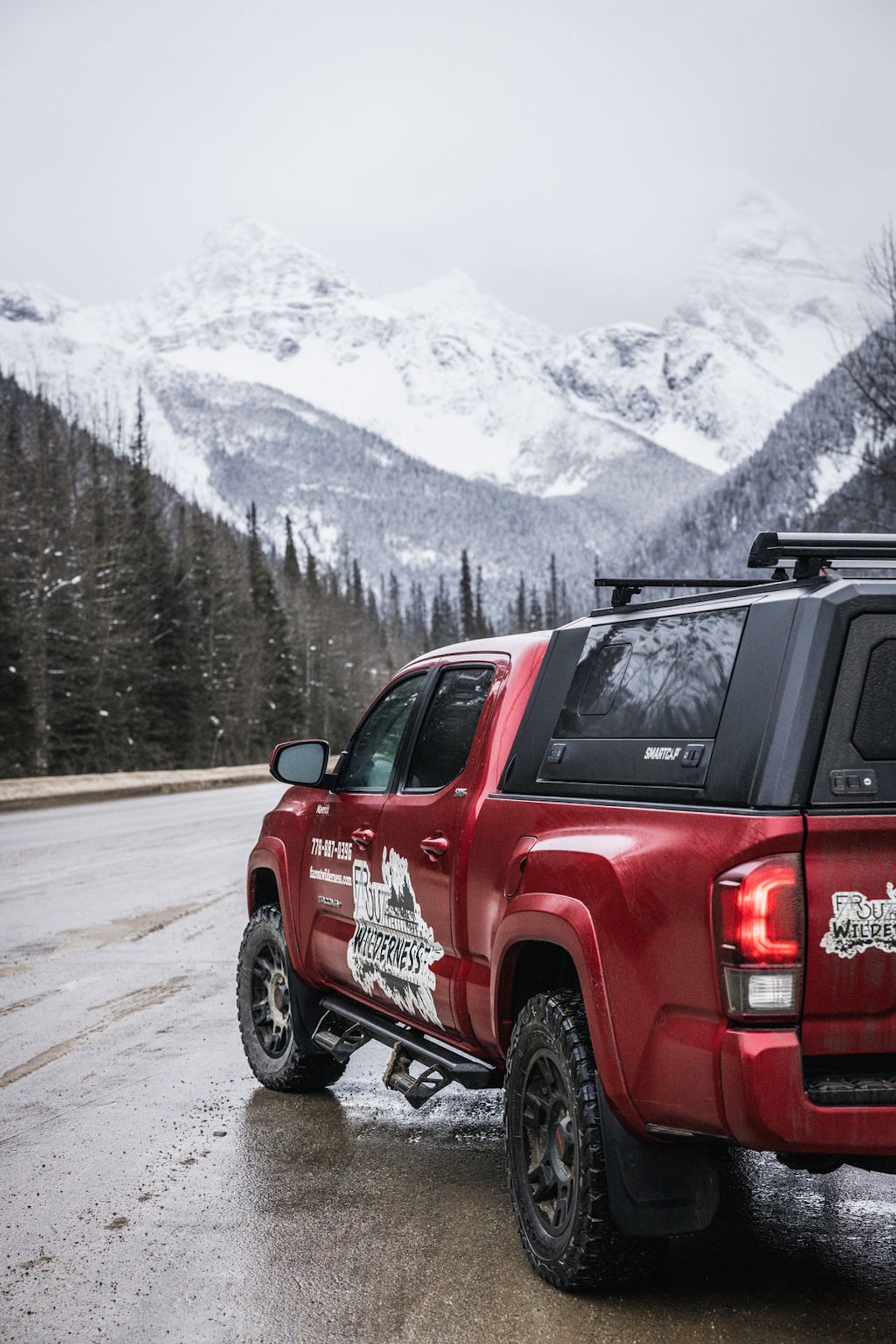 Red 4x4 truck parked on a snowy mountain road with forest trees and snow‑covered peaks.