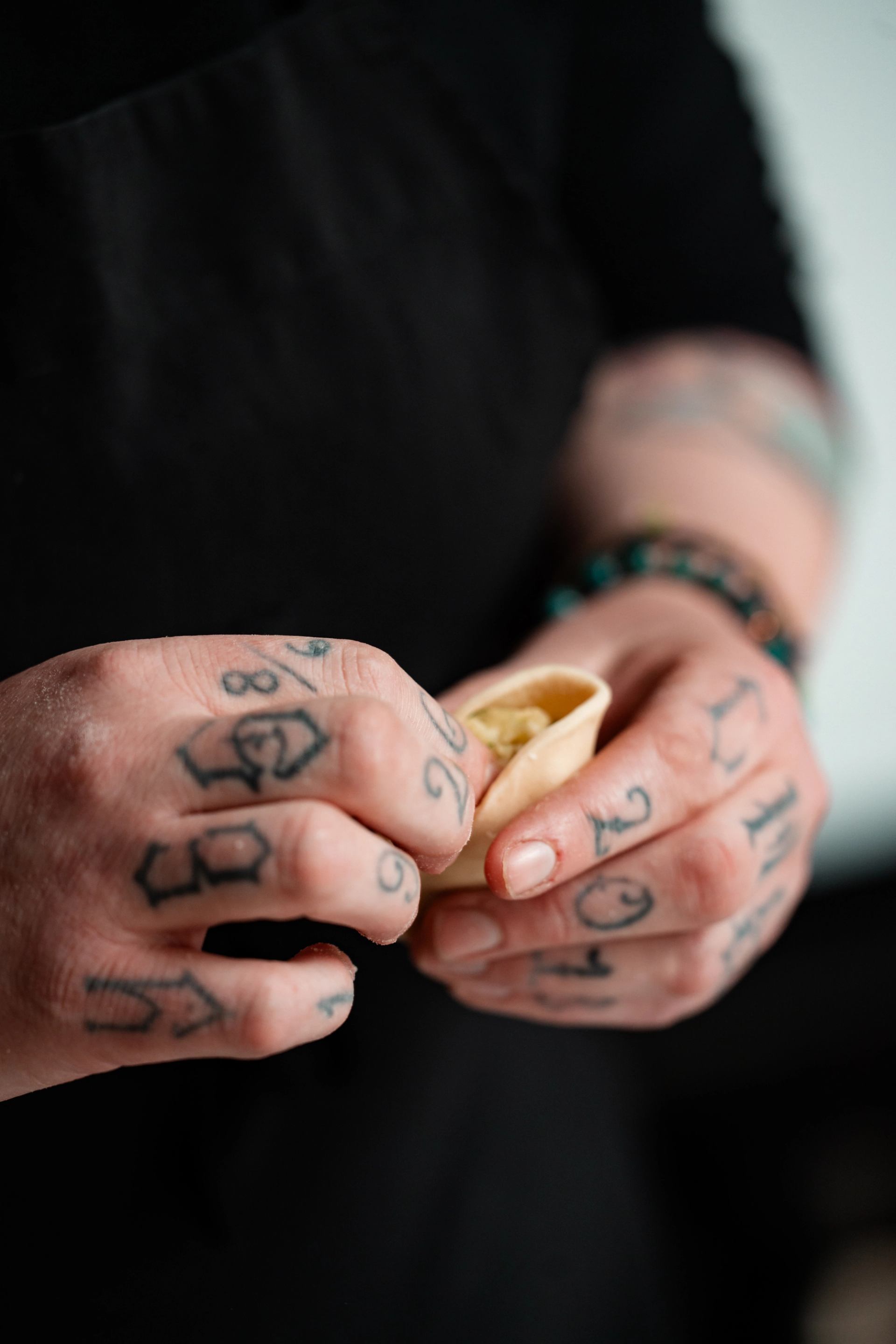 Tattooed hands in black apron shaping a small dumpling with filling.