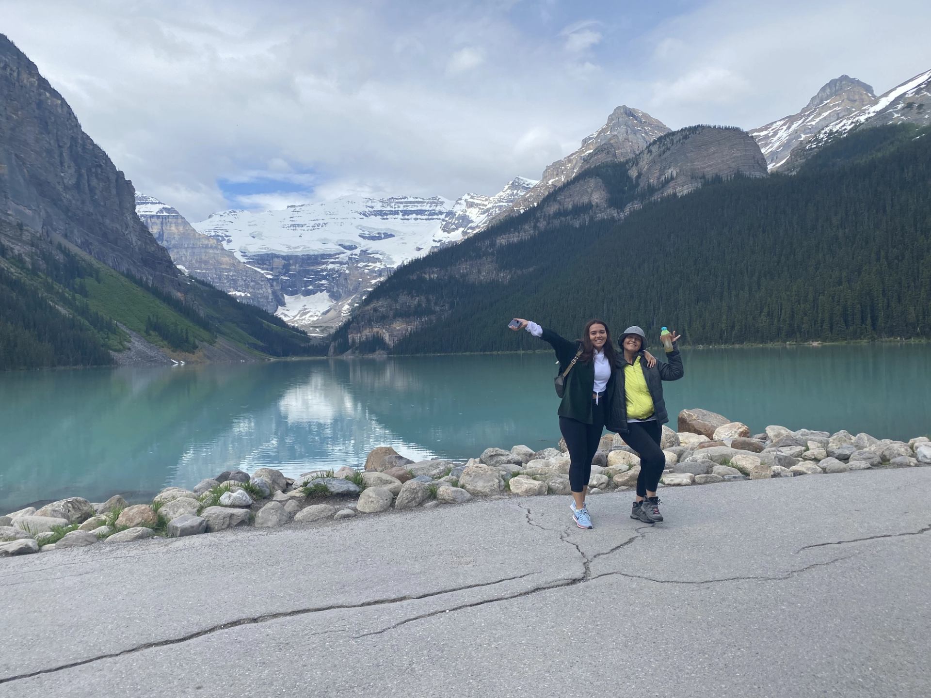 Two people pose on lakeside road with turquoise water and snow-capped mountain backdrop.