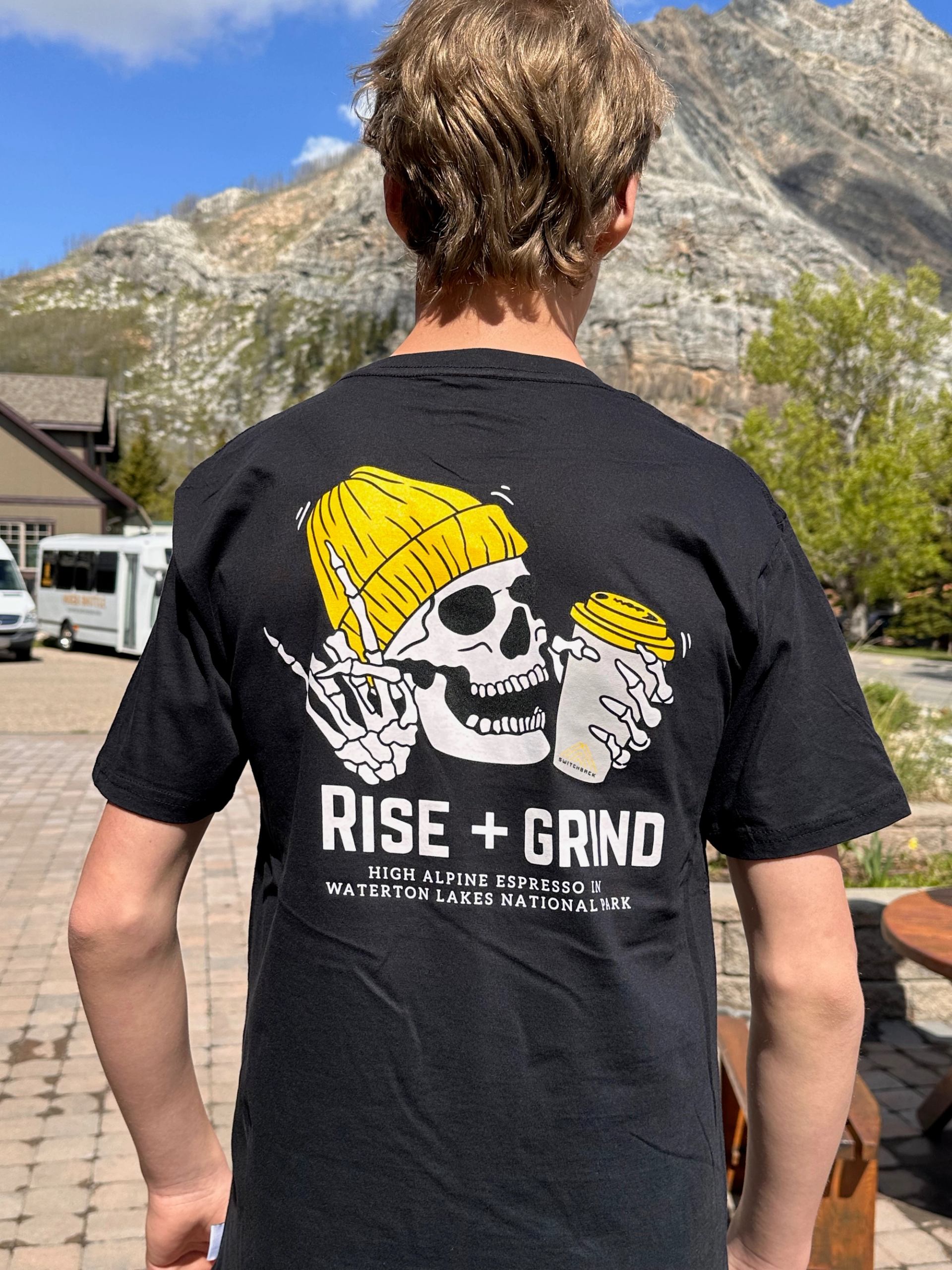 Person wearing a Rise + Grind Switchback T‑shirt with mountains and café buildings behind.