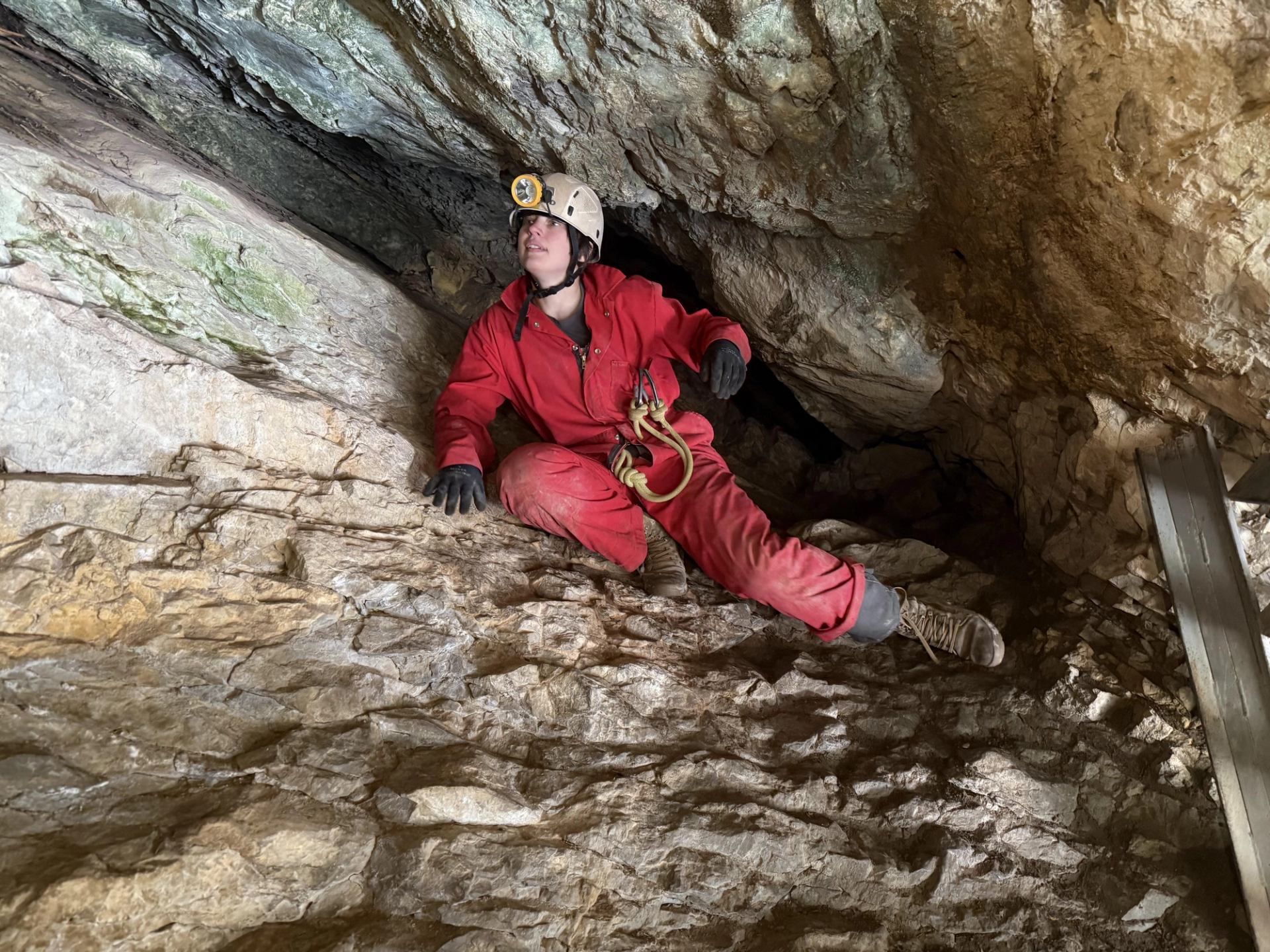 Caver in red coveralls and a headlamp sits on a rocky cave ledge.