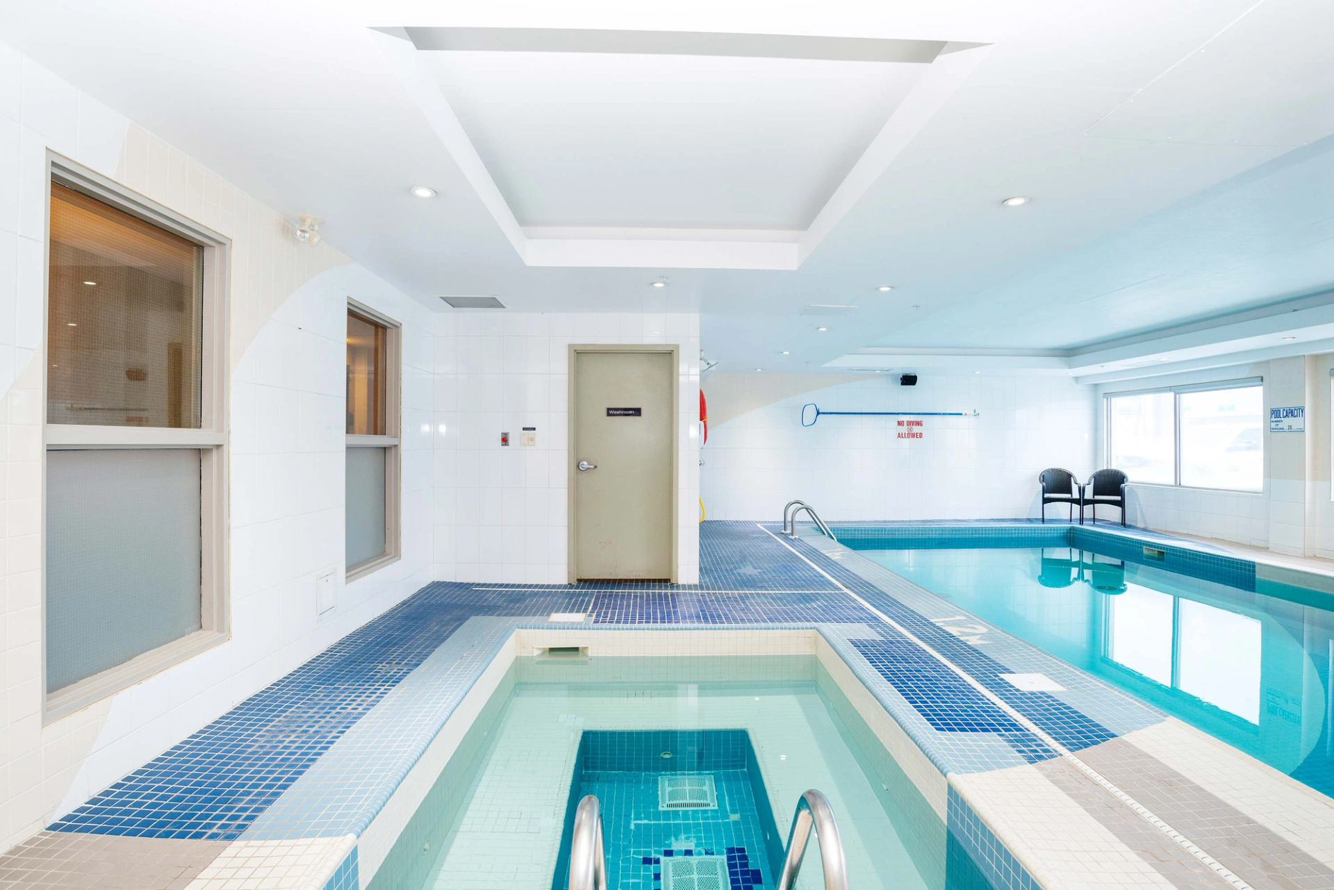 Indoor hotel pool area with hot tub, lap pool, tiled deck and windows.