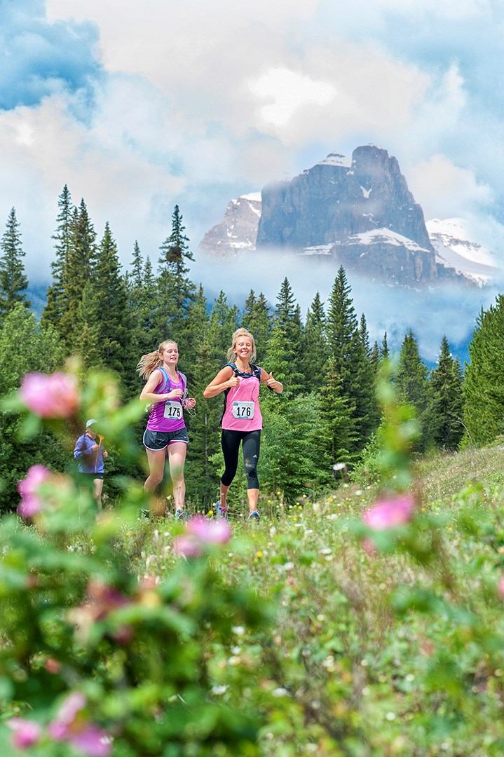 Two runners on forest trail with mountain and flowers.