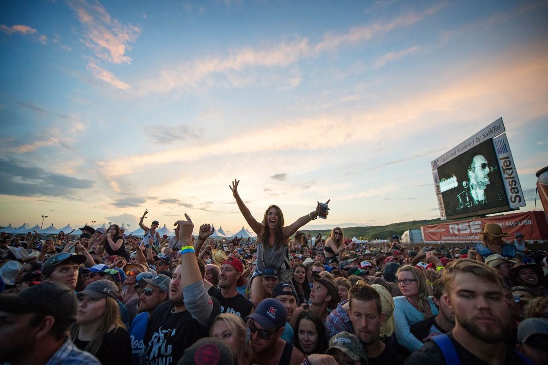 Concert crowd under colourful sky with person raised above others and large video screen.