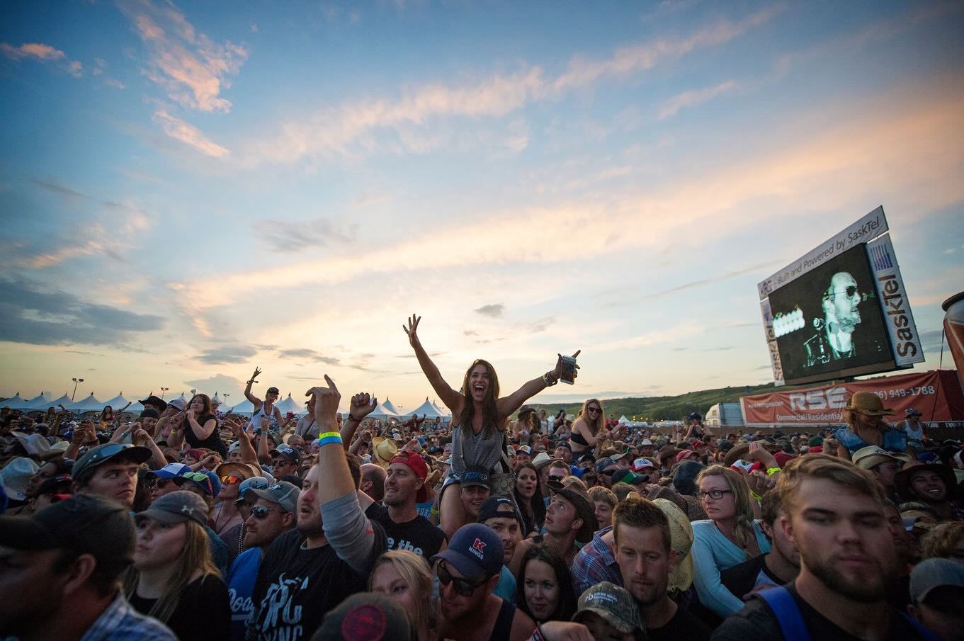 Concert crowd under colourful sky with person raised above others and large video screen.