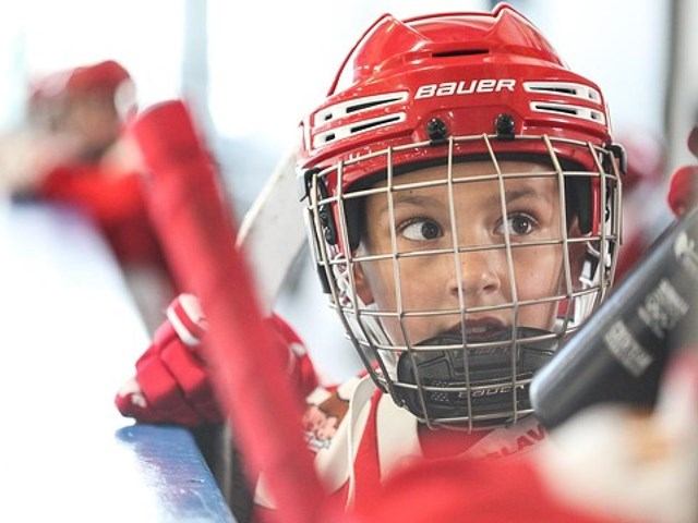 Hockey player in red helmet and cage leans on rink boards.