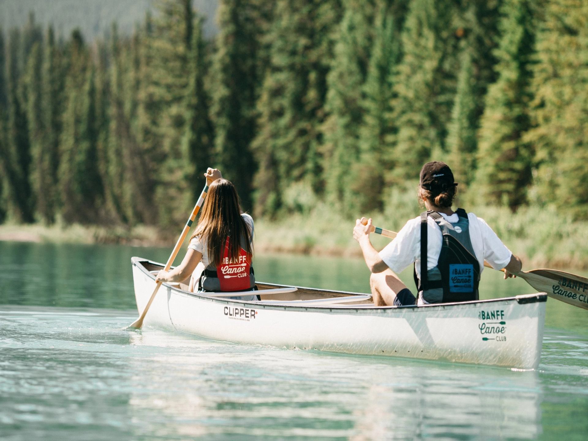 Two people paddling a canoe on calm water with a forested shoreline in the background.