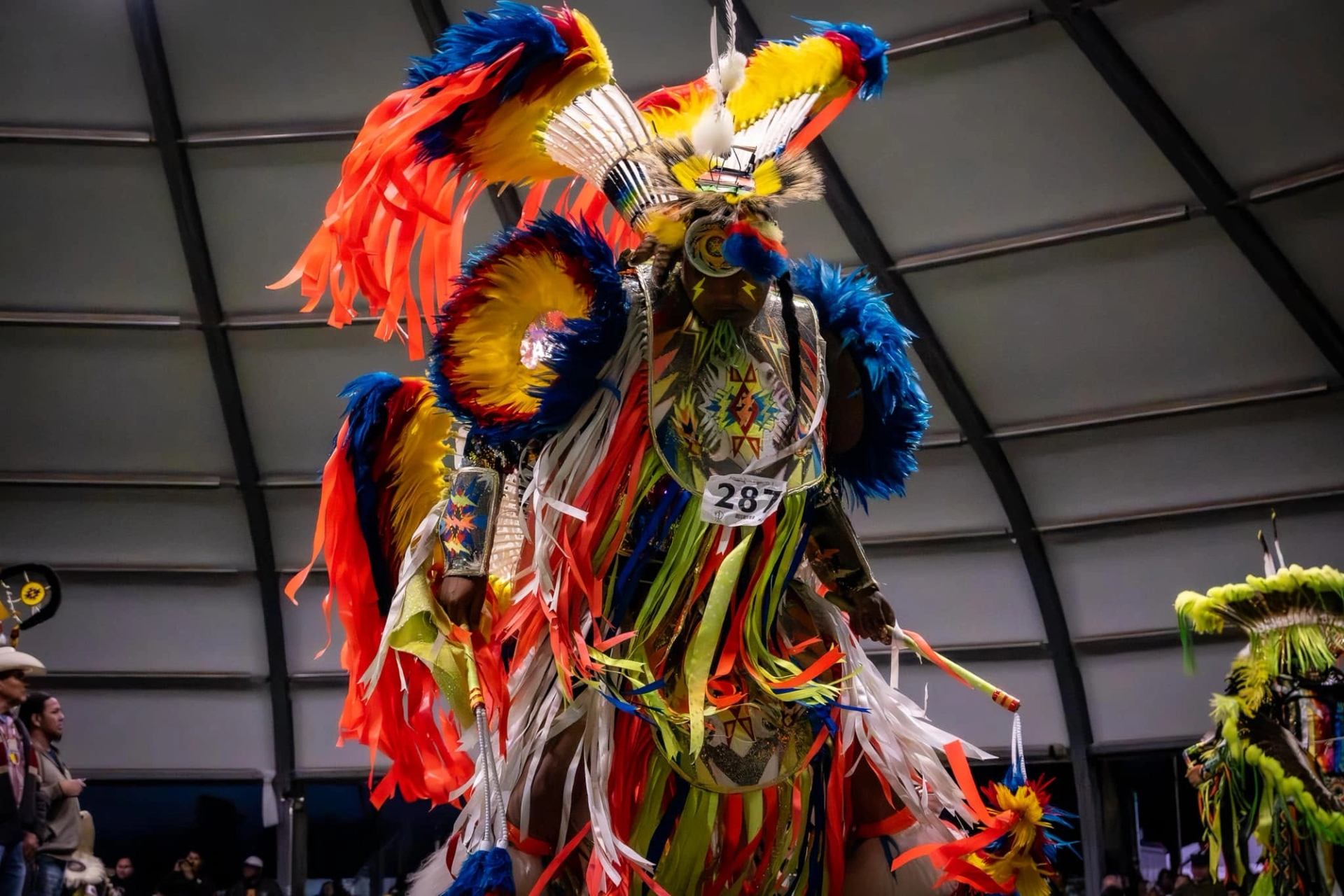 Powwow dancer wearing bright feathered regalia and ribbons performs inside large arena.