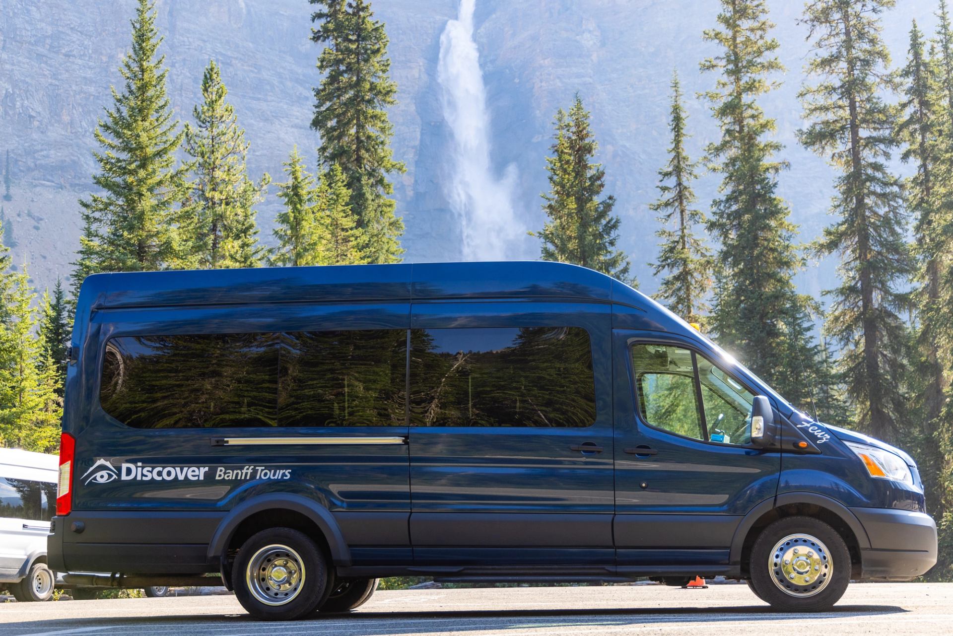 Blue Discover Banff Tours van parked on road with tall trees and waterfall behind.