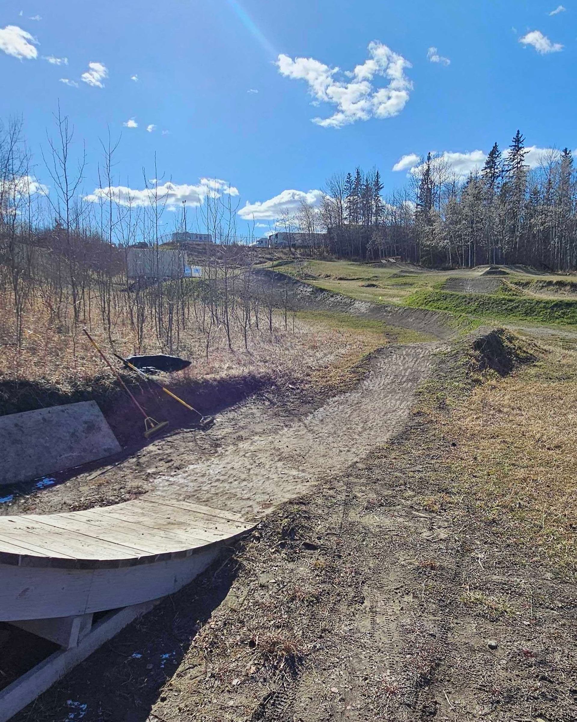 Dirt bike trail with wooden ramp, grassy mounds and sparse trees under blue sky.