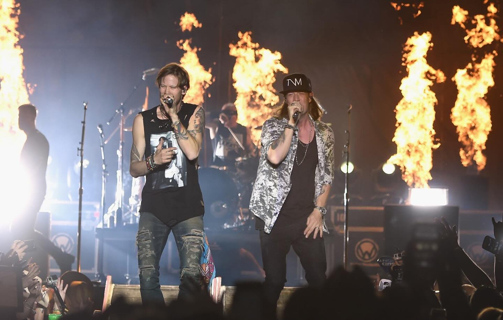 Two singers perform onstage with microphones as flames rise behind them and crowd watches.