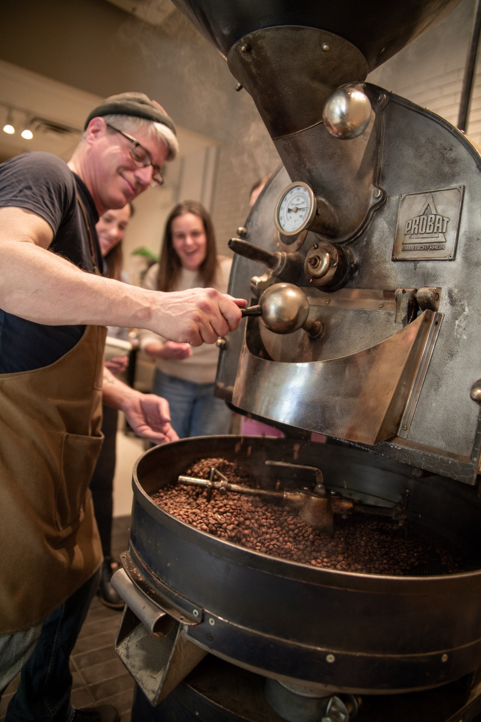 Coffee roaster operates large Probat machine while people watch beans tumbling inside drum.