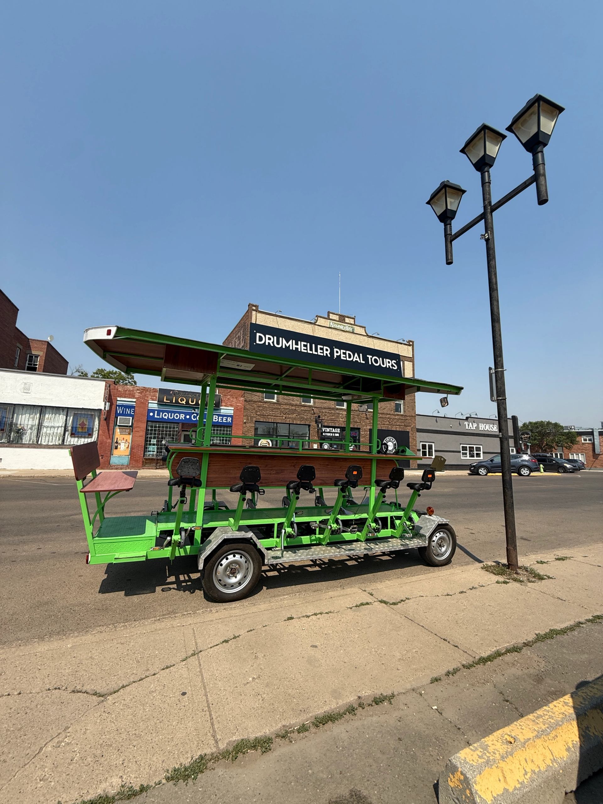 Green pedal tour cart in front of Drummheller Pedal Tours storefront sign, streetlights visible.