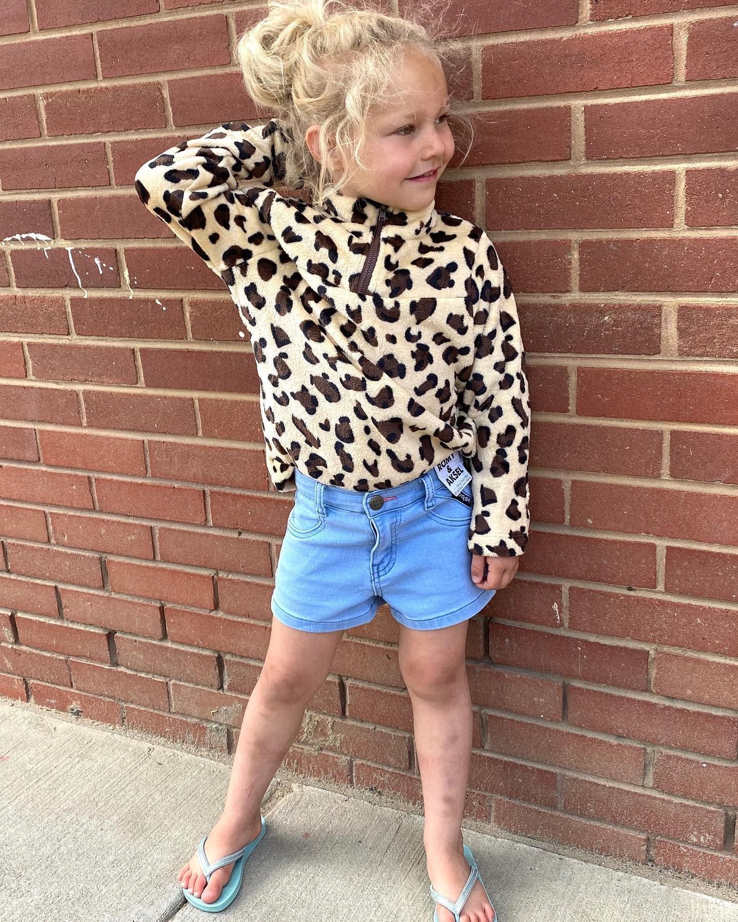 Girl in leopard-print jacket and denim shorts standing in front of brick wall.