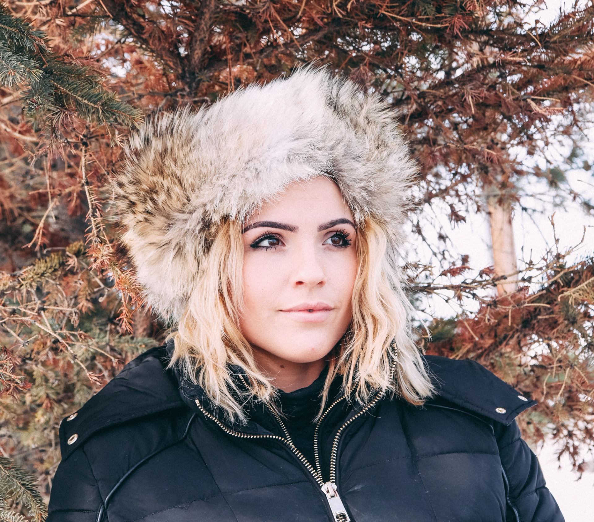 Woman in fur-trimmed hat and black winter coat stands outdoors near pine trees, looking thoughtfully.