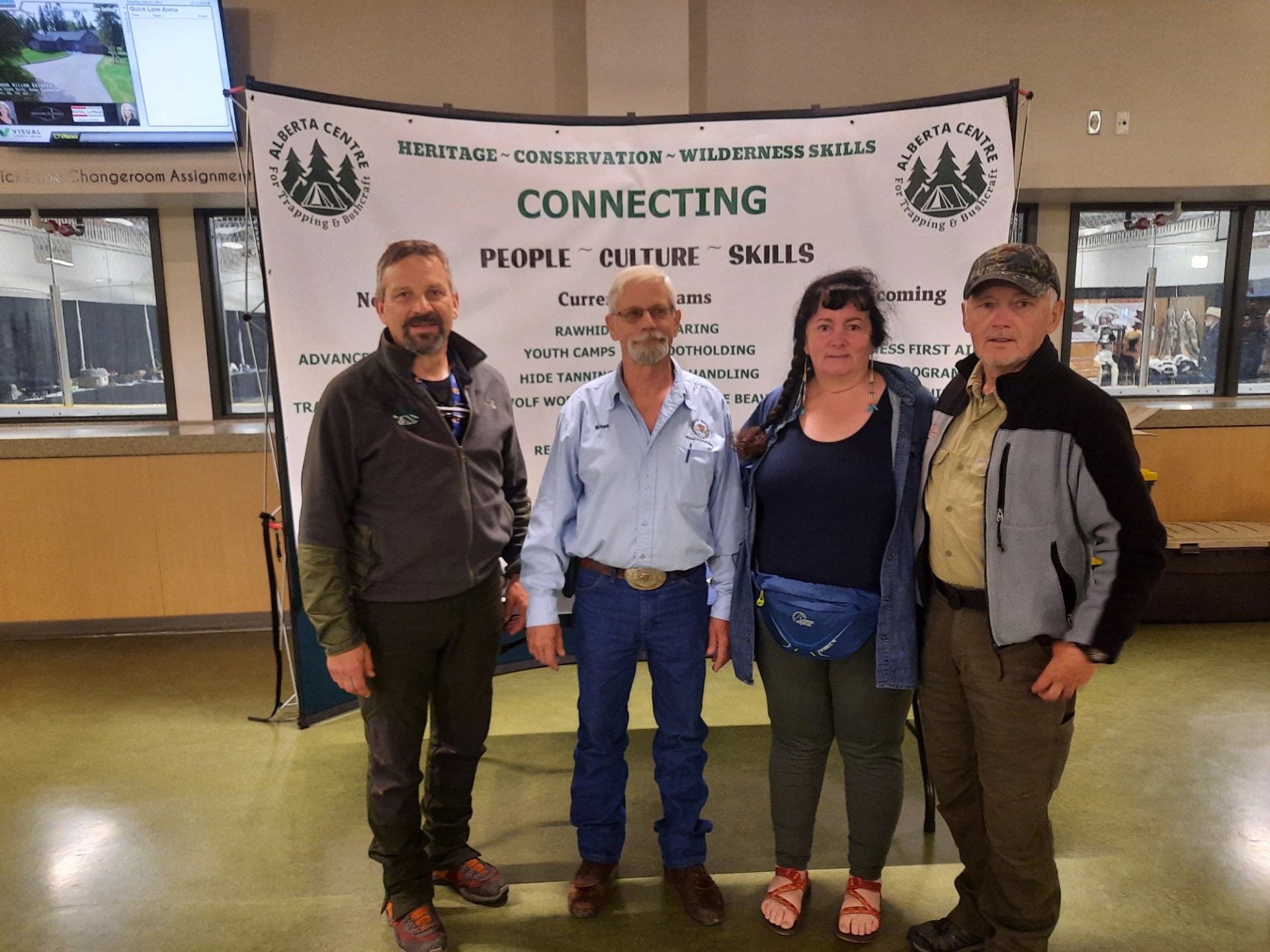 Four adults stand in front of Alberta Centre trapping and bushcraft information banner indoors.