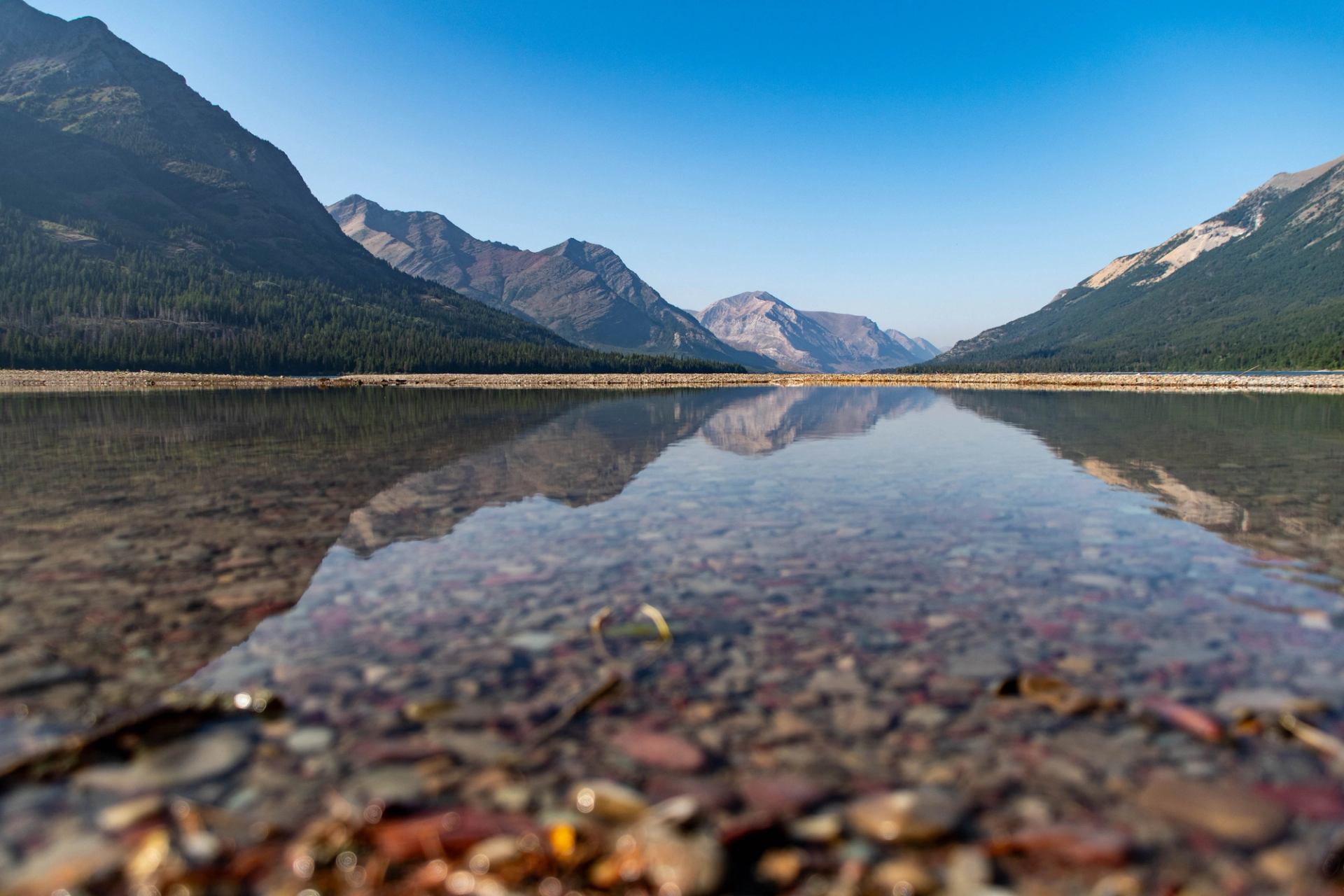 Mountain valley and lake with reflections, forested slopes, and rocky shoreline in foreground.