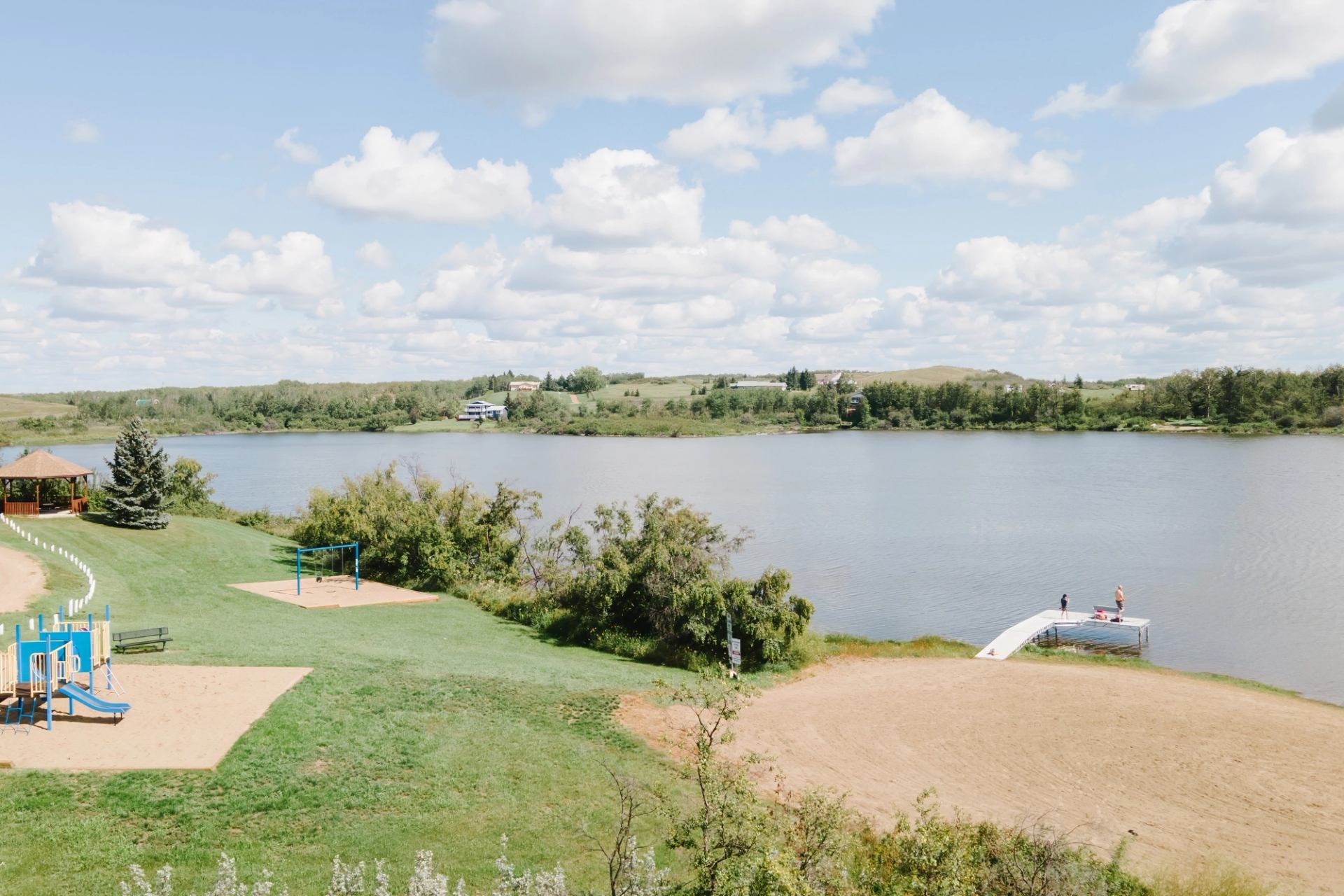 Lakeside park with playground, sandy beach, dock and people standing near calm water.