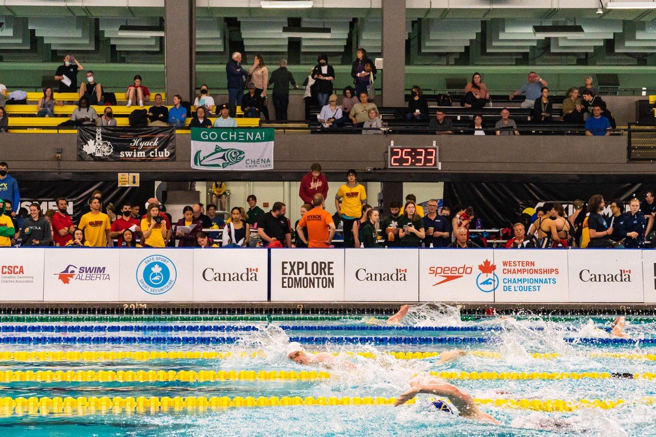 Competitive swimmers race in indoor pool as spectators watch under Western Championships banners.