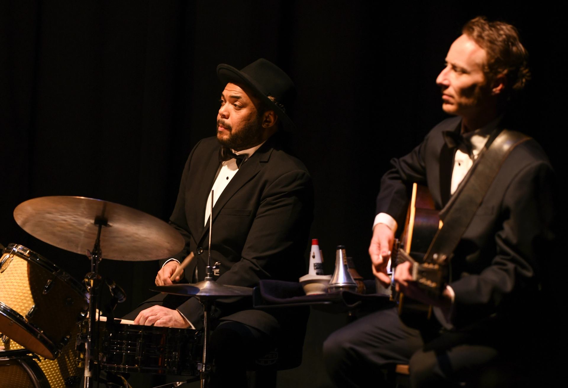 Jazz drummer and guitarist in tuxedos perform on stage with dark background.