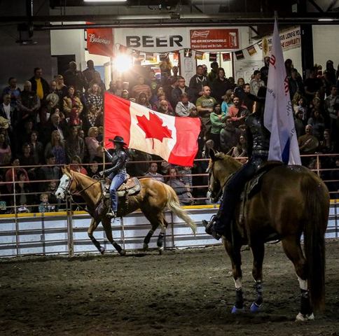 Riders on horseback carrying flags inside a rodeo arena with a large crowd watching.