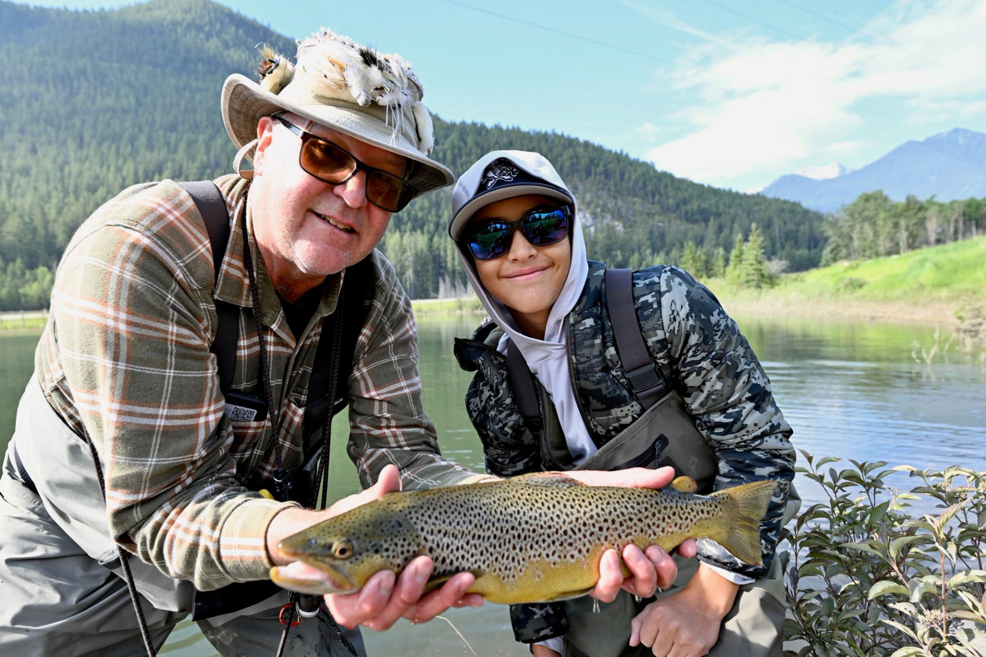 Two anglers in waders hold a large brown trout beside a forested lake.