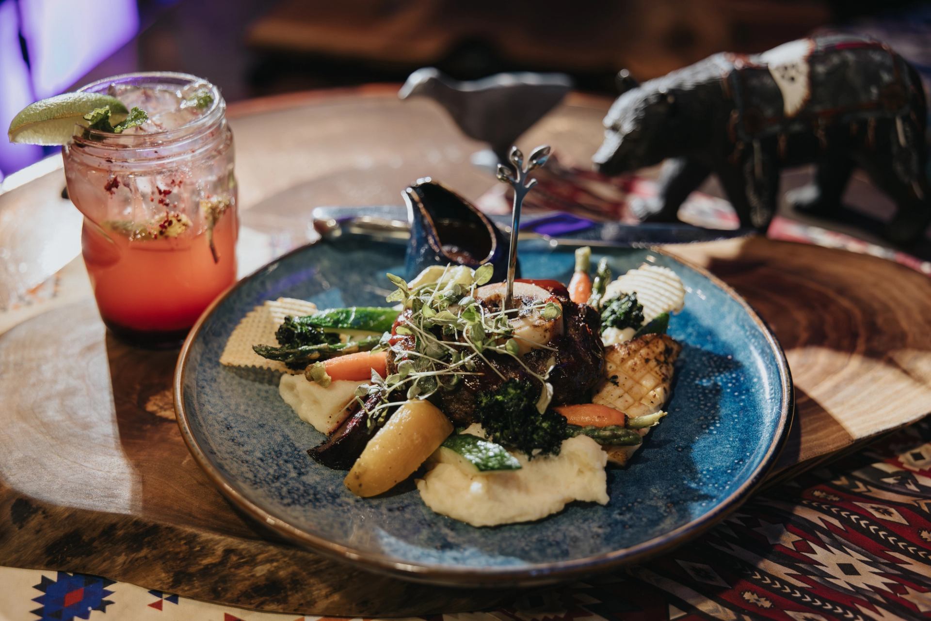 Blue plate with meat, mashed potatoes and vegetables beside pink cocktail on wooden table.