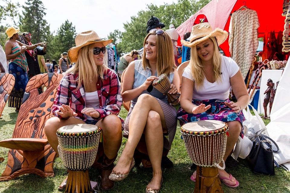 Women sitting outdoors and playing hand drums at a cultural festival booth.