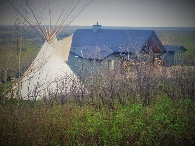 A tipi stands beside a modern house surrounded by leafless trees and green shrubs under an overcast sky, blending cultural heritage and contemporary living.