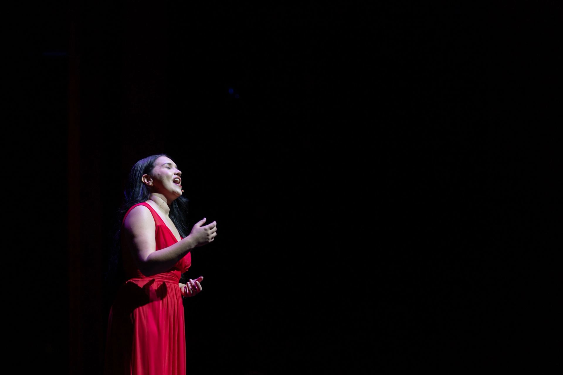 Performer in red dress under spotlight on dark stage during opera show.