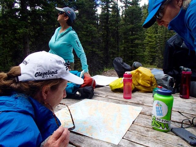 Three women studying a map at a wooden picnic table in a forested area, surrounded by hiking gear and water bottles, with trees in the background.