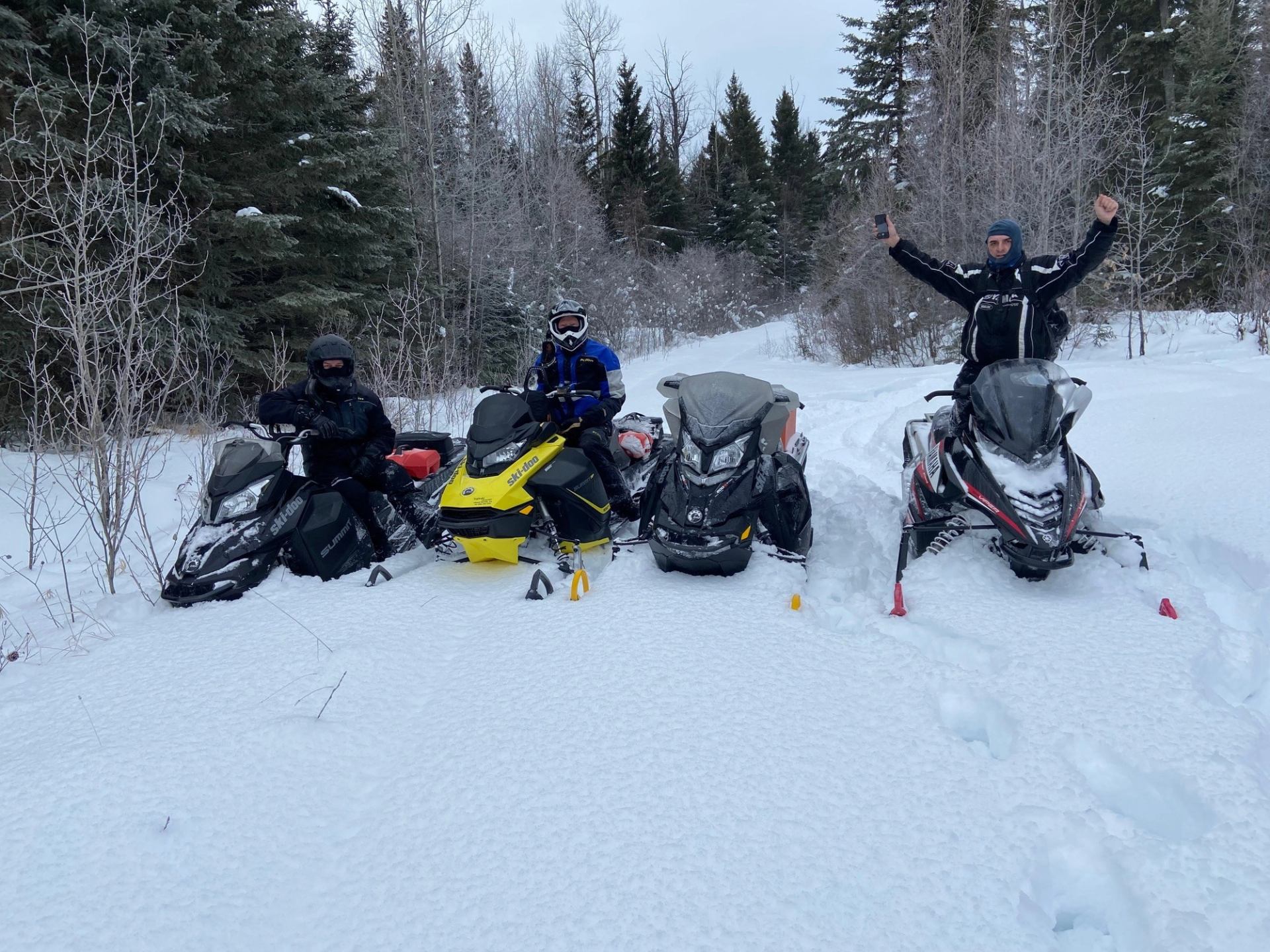 Four people on snowmobiles in snowy forest; three wear helmets, one raises arms, showing adventure and joy.