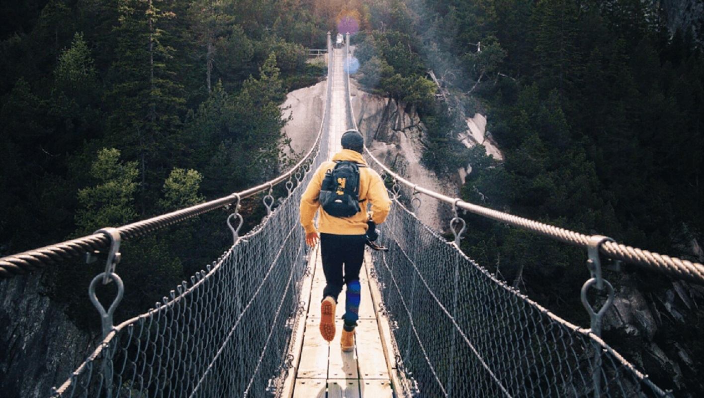 Hiker walking across a long suspension bridge high above a forested mountain canyon.