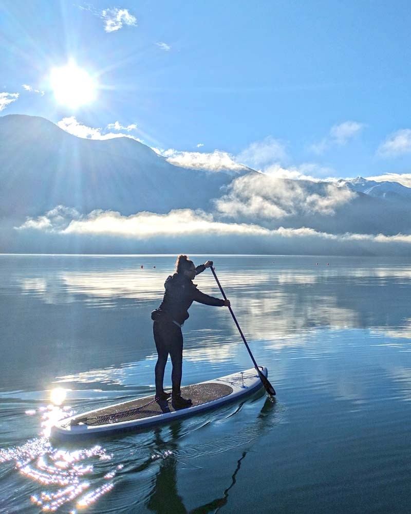 Person paddleboarding on calm water with mountain backdrop and bright sun.