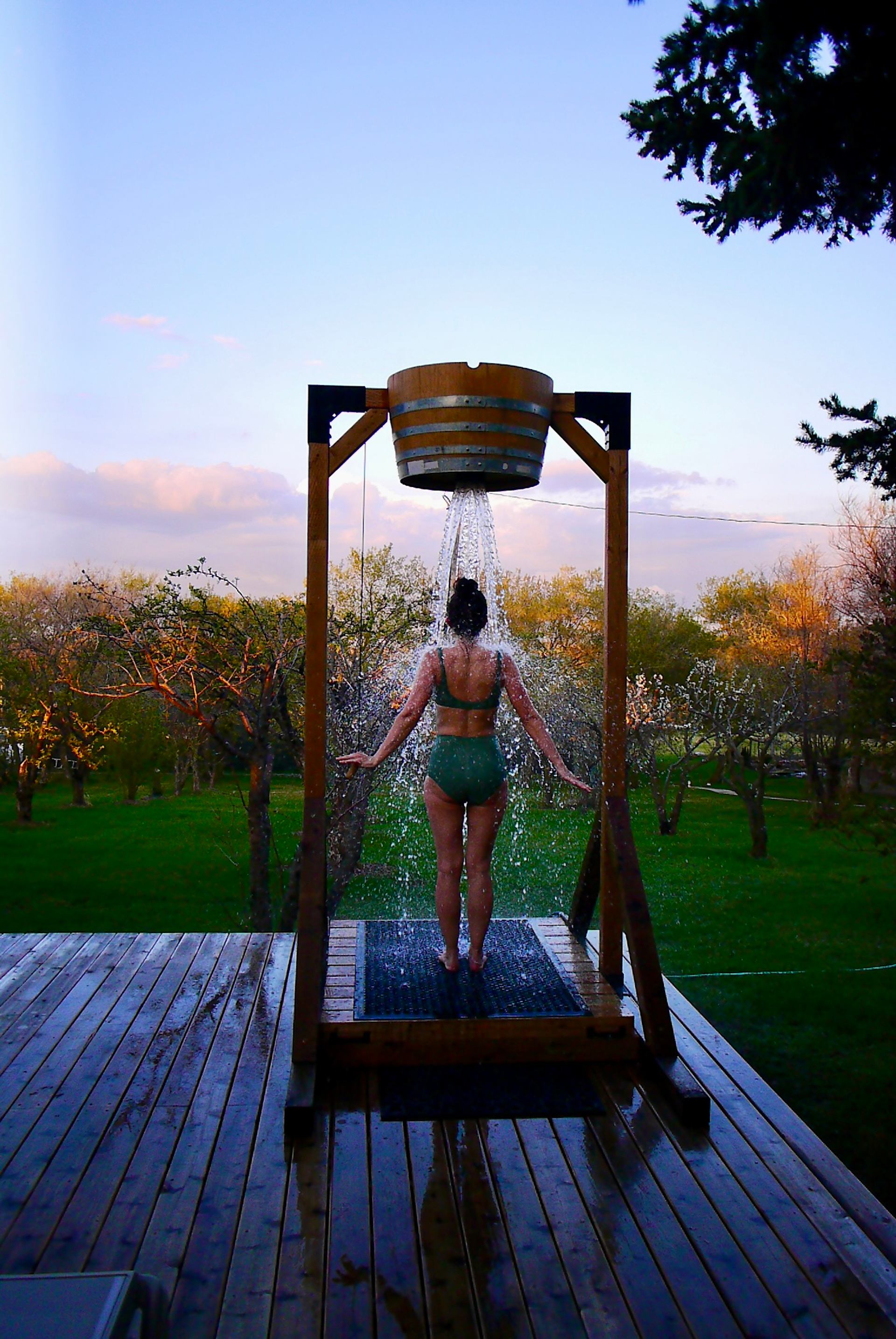 A person stands beneath a large wooden bucket dumping water overhead on an outdoor platform. The scene overlooks a grassy area with trees under a soft evening sky.
