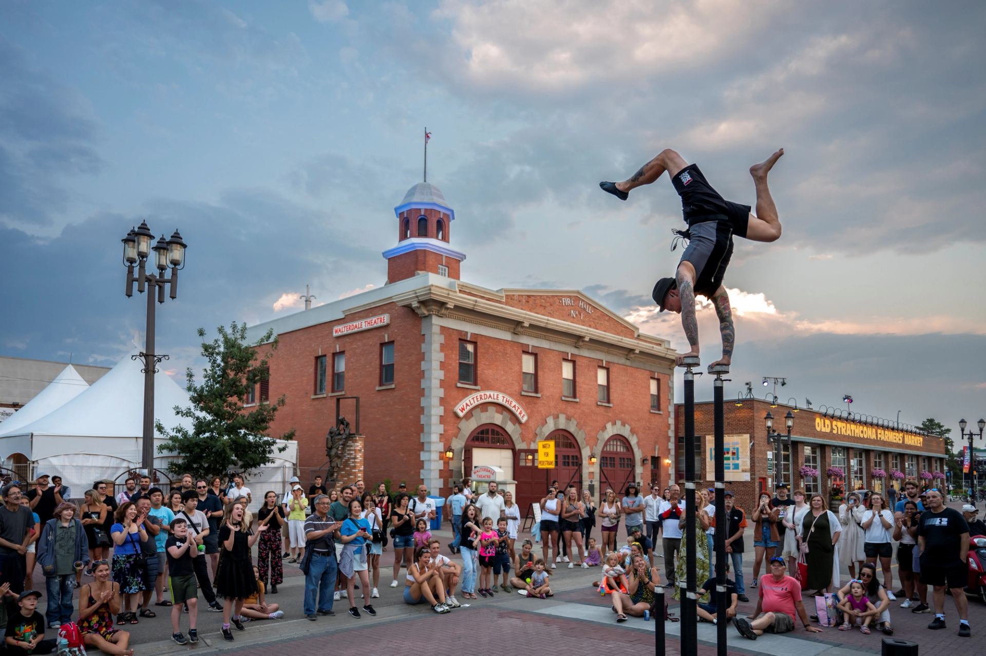 A street performer balances on their hands atop tall poles, entertaining a crowd in front of historic brick buildings.