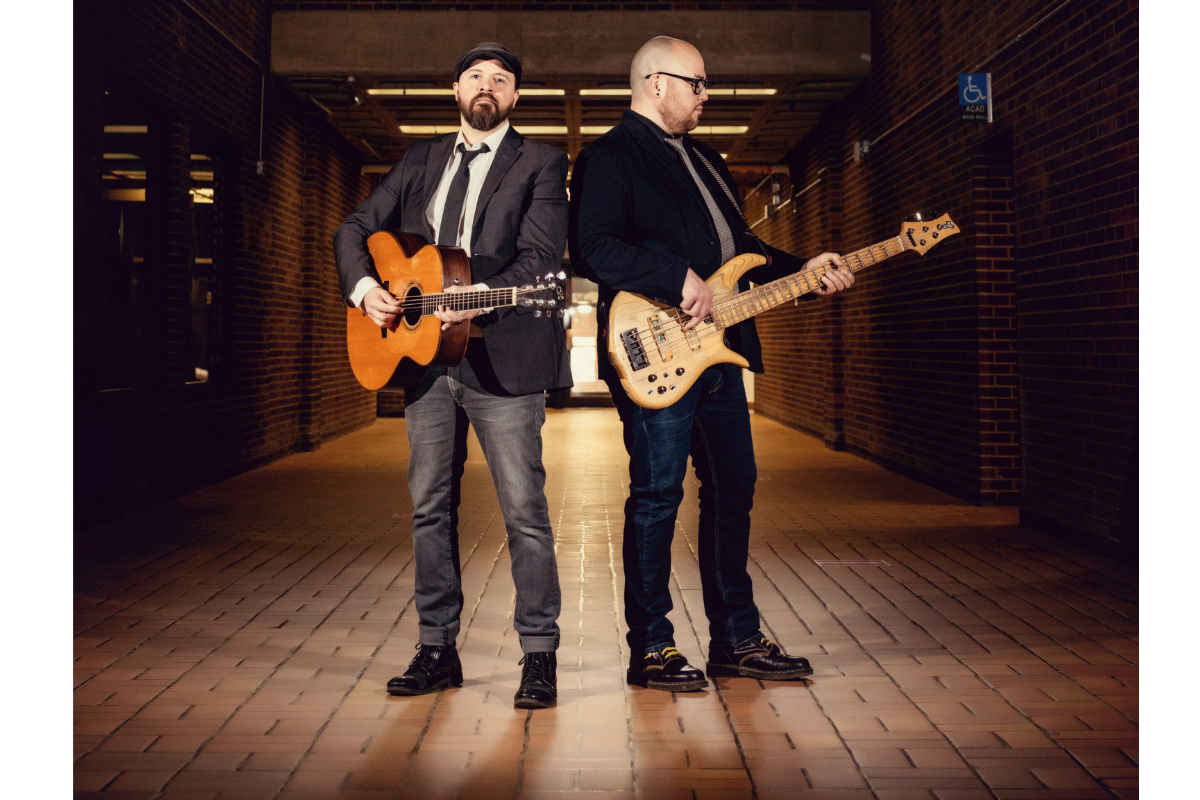 Musicians hold an acoustic guitar and electric bass in a brick hallway.