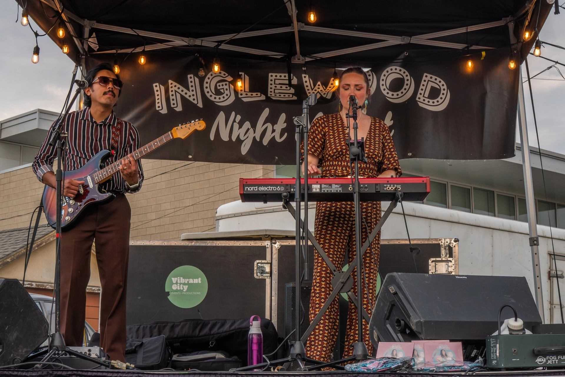 Musicians perform on outdoor stage with guitar and keyboard under Inglewood Night Market banner.
