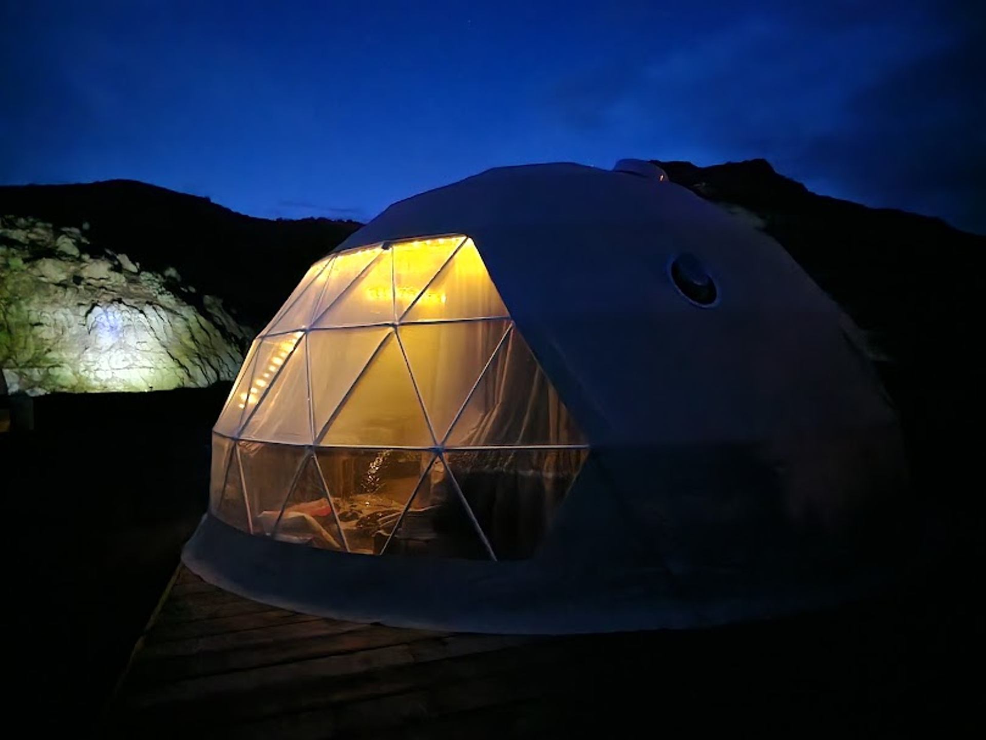 Geodesic dome tent glows with warm interior light against dark rocky hillside and sky.