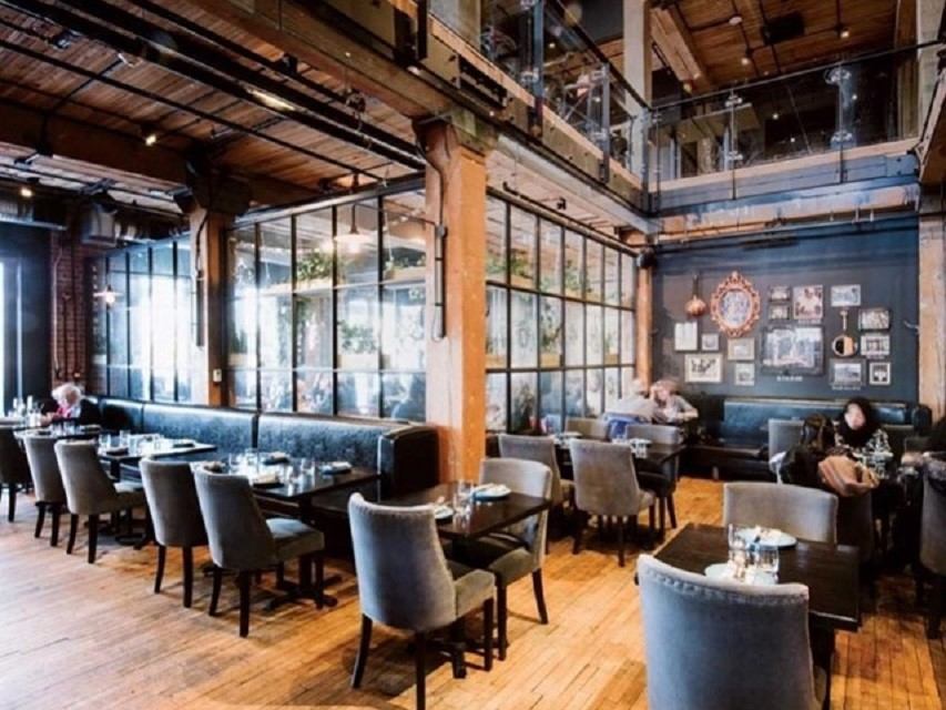 Spacious, rustic restaurant with wooden floors, exposed beams, gray upholstered chairs, and glass partition. Cozy atmosphere with natural light.