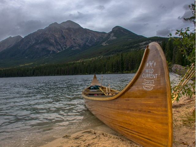 A wooden canoe labeled "Wild Current" rests on a sandy lakeshore, with a backdrop of towering, cloudy mountains and a dense forest, creating a tranquil and adventurous tone.