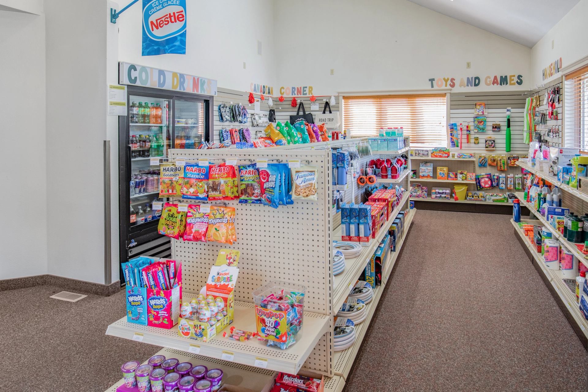 A bright camp store offers snacks, cold drinks, games, toys, and travel essentials on display shelves.