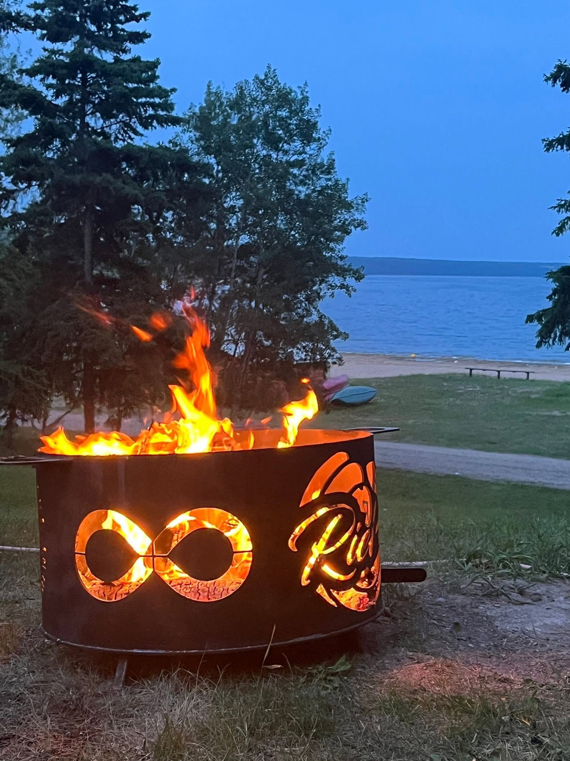 Metal fire pit with infinity symbol burns near lakeshore, trees and sandy beach.