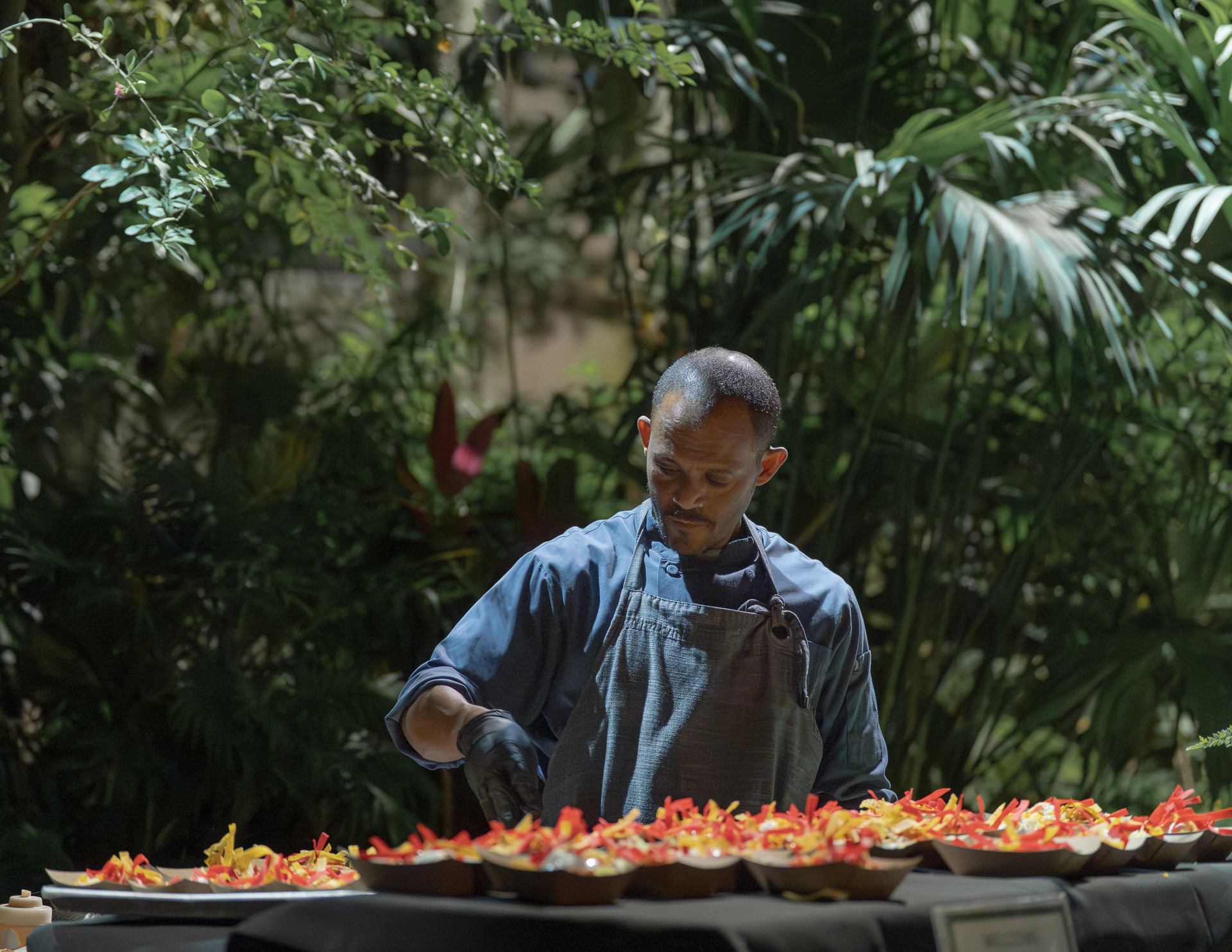 Chef prepares colourful plated dishes at an outdoor table surrounded by tropical plants.