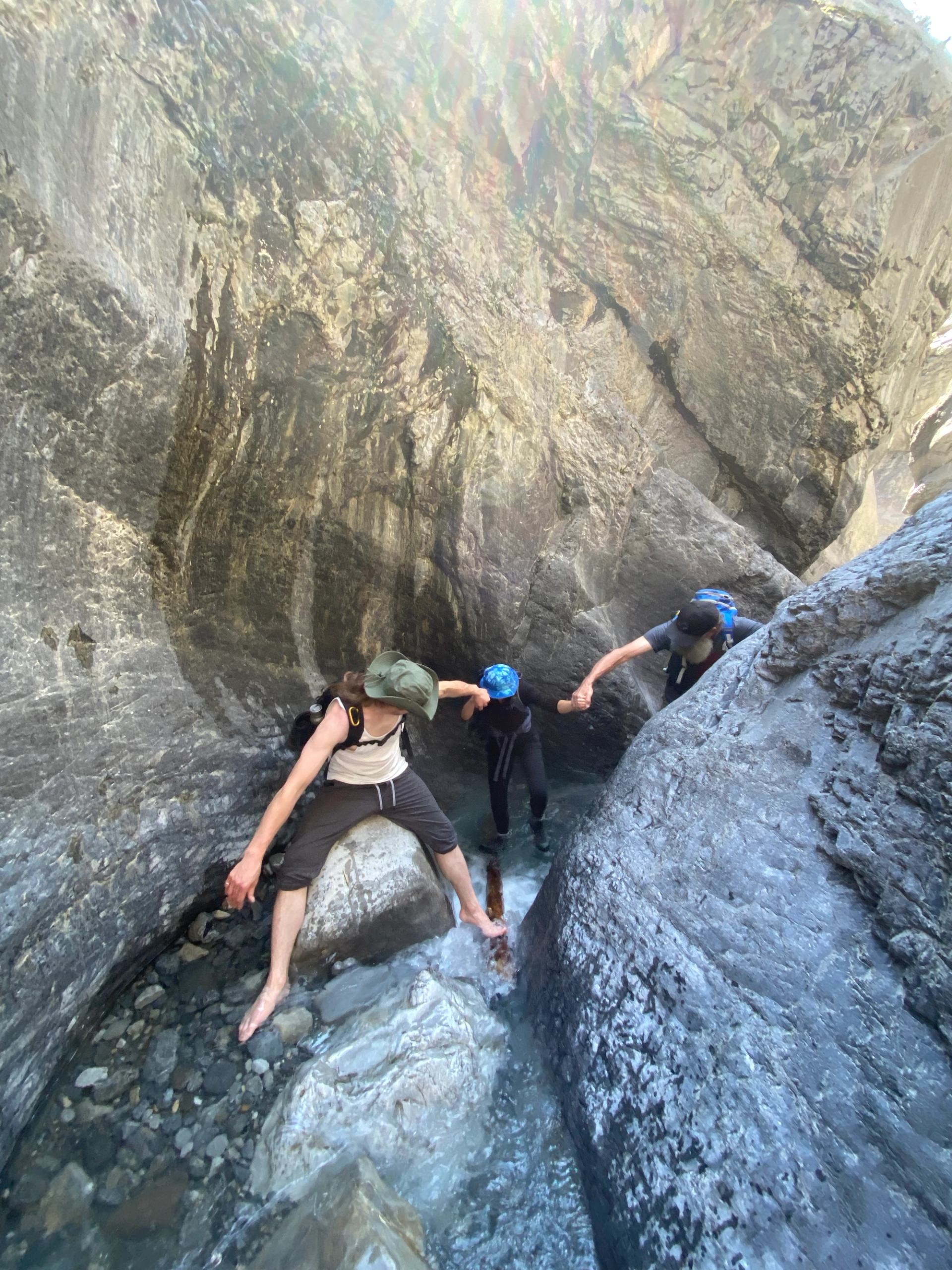 Three hikers wade through narrow rocky canyon, holding hands above shallow flowing water.