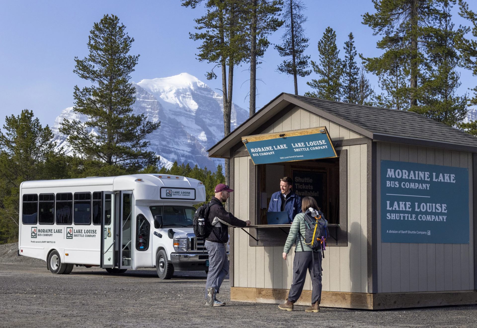 Shuttle ticket kiosk and parked Moraine Lake bus with hikers, trees and snowy mountain backdrop.