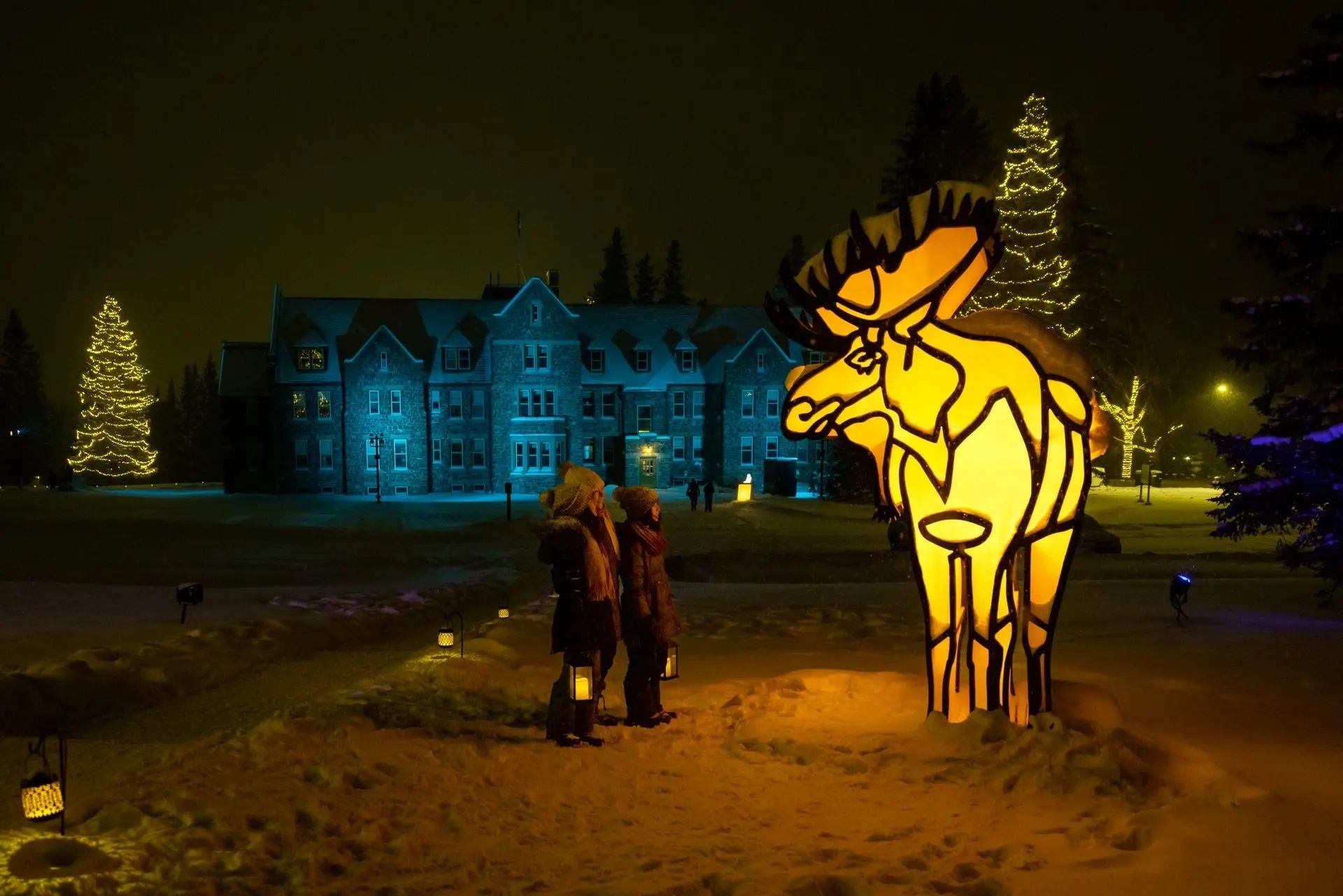 Three people admire a glowing moose sculpture in a snowy, festive scene with lit-up buildings.