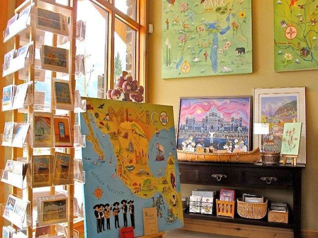 Interior gallery corner with colorful maps, framed art, and a postcard display rack.