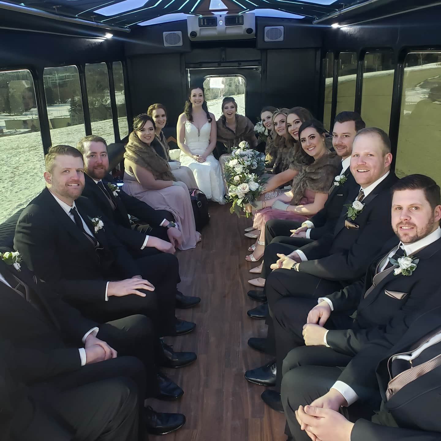 Group in formal attire seated inside a bright, wood-floored party bus.